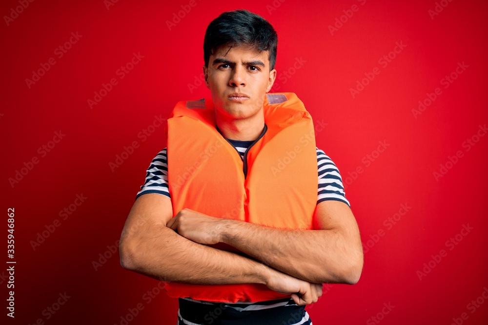 Young handsome man wearing orange safety life jacket over isolated red background skeptic and nervous, disapproving expression on face with crossed arms. Negative person. Young handsome man wearing orange safety life jacket over isolated red background skeptic and nervous, disapproving expression on face with crossed arms. Negative person.