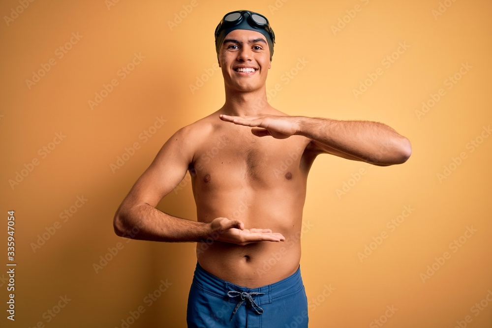 Young handsome man shirtless wearing swimsuit and swim cap over isolated yellow background gesturing with hands showing big and large size sign, measure symbol. Smiling looking at the camera. Measure