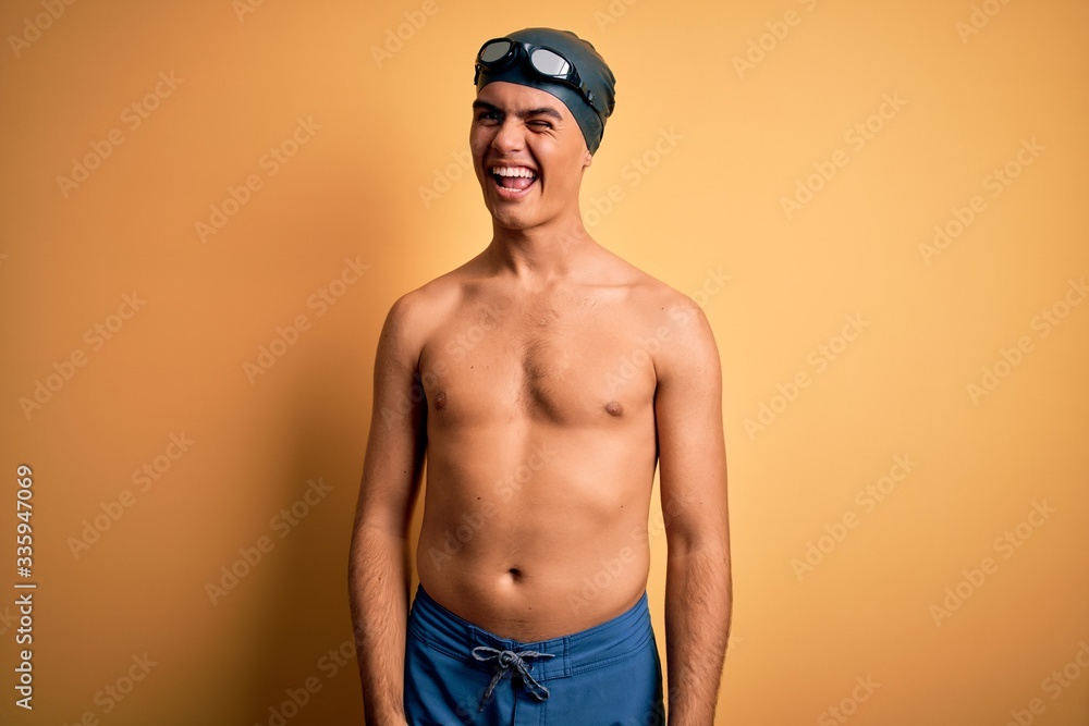 Young handsome man shirtless wearing swimsuit and swim cap over isolated yellow background winking looking at the camera with sexy expression, cheerful and happy face.