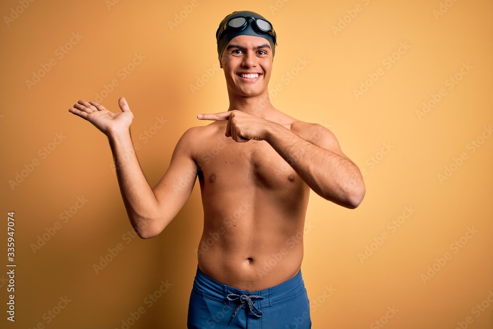 Young handsome man shirtless wearing swimsuit and swim cap over isolated yellow background amazed and smiling to the camera while presenting with hand and pointing with finger.