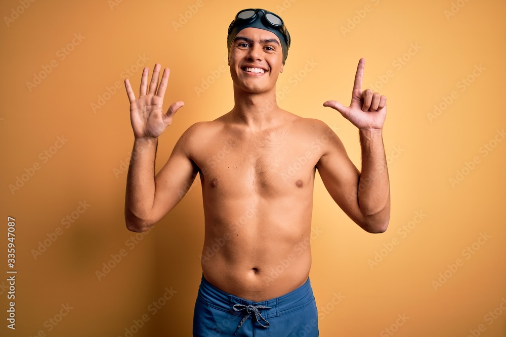 Young handsome man shirtless wearing swimsuit and swim cap over isolated yellow background showing and pointing up with fingers number seven while smiling confident and happy. Young handsome man shirtless wearing swimsuit and swim cap over isolated yellow background showing and pointing up with fingers number seven while smiling confident and happy.