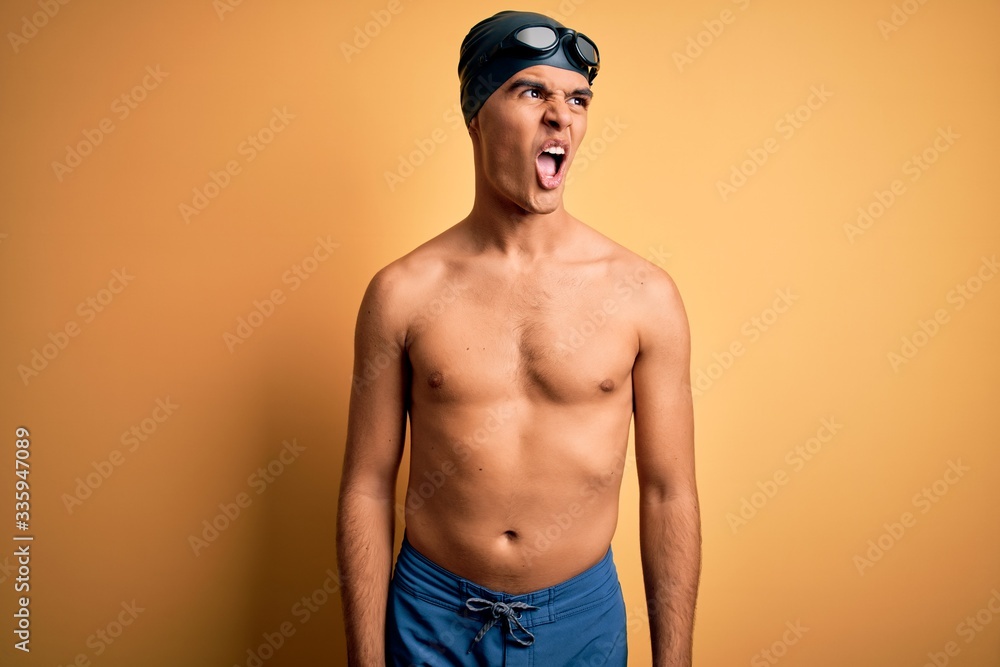 Young handsome man shirtless wearing swimsuit and swim cap over isolated yellow background angry and mad screaming frustrated and furious, shouting with anger. Rage and aggressive concept.