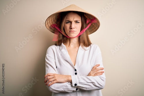 Young beautiful redhead woman wearing asian traditional conical hat over white background skeptic and nervous, disapproving expression on face with crossed arms. Negative person.