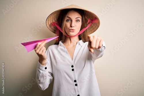 Young beautiful redhead woman wearing asian traditional hat holding paper airplane pointing with finger to the camera and to you, hand sign, positive and confident gesture from the front
