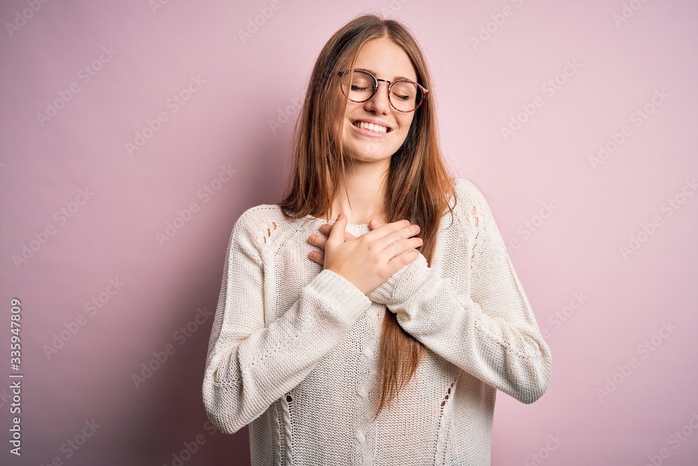 Young beautiful redhead woman wearing casual sweater and glasses over pink background smiling with hands on chest with closed eyes and grateful gesture on face. Health concept. Young beautiful redhead woman wearing casual sweater and glasses over pink background smiling with hands on chest with closed eyes and grateful gesture on face. Health concept.