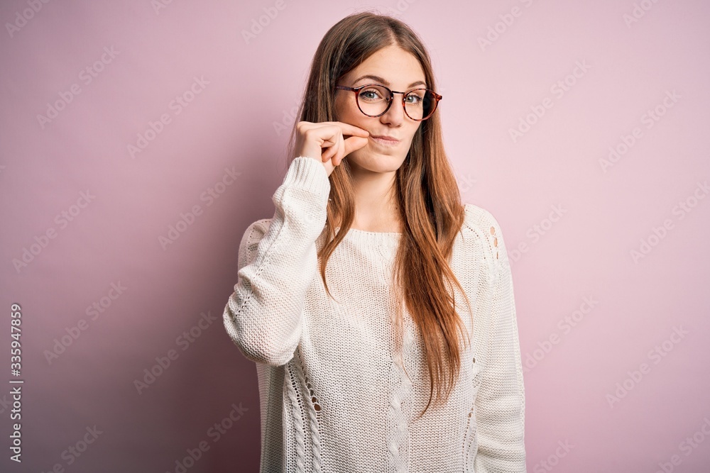 Young beautiful redhead woman wearing casual sweater and glasses over pink background mouth and lips shut as zip with fingers. Secret and silent, taboo talking