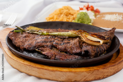 A view of a carne asada skillet plate, with a side of rice and beans, in a restaurant or kitchen setting.