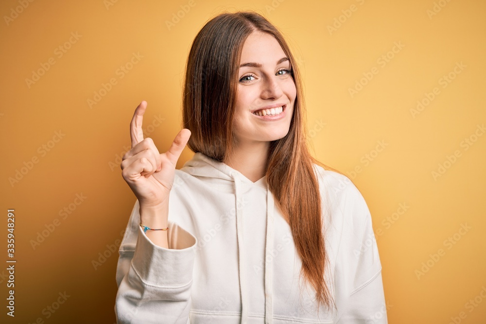 Young beautiful redhead sporty woman wearing sweatshirt over isolated yellow background smiling and confident gesturing with hand doing small size sign with fingers looking and the camera. Measure Young beautiful redhead sporty woman wearing sweatshirt over isolated yellow background smiling and confident gesturing with hand doing small size sign with fingers looking and the camera. Measure