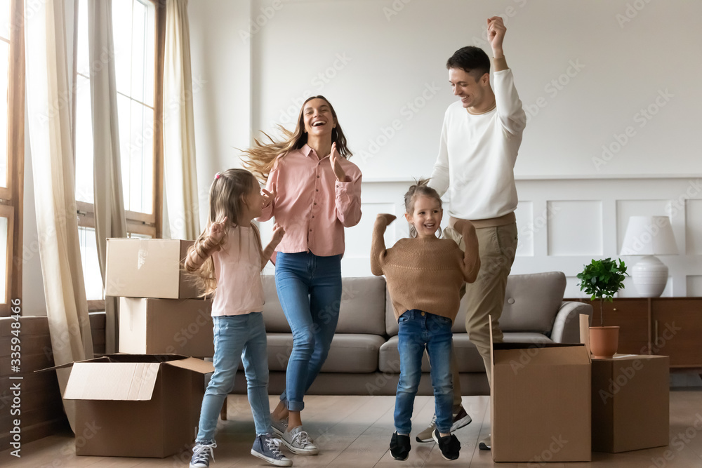 Full length overjoyed young family couple having fun with adorable two daughters after moving in new apartment. Happy parents dancing with cute children among big cardboard boxes in living room. Full length overjoyed young family couple having fun with adorable two daughters after moving in new apartment. Happy parents dancing with cute children among big cardboard boxes in living room.