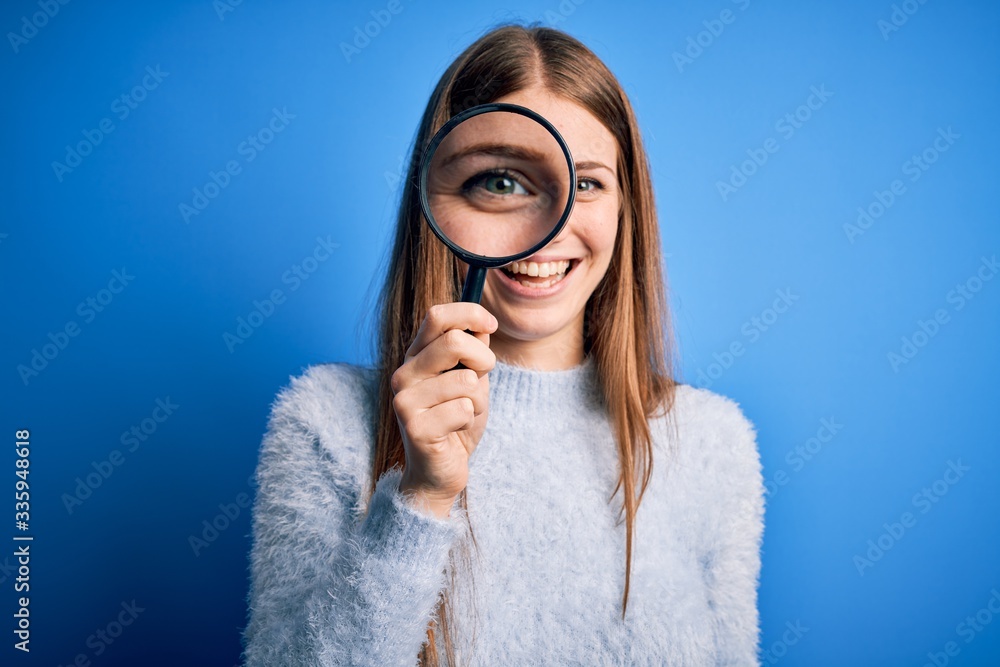 Young beautiful redhead detective woman using magnifying glass over isolated blue background with a happy face standing and smiling with a confident smile showing teeth Young beautiful redhead detective woman using magnifying glass over isolated blue background with a happy face standing and smiling with a confident smile showing teeth