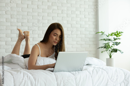 Beautiful asian woman working on a laptop sitting on the bed in the house.Woman checking social apps and working.Work from home concept.