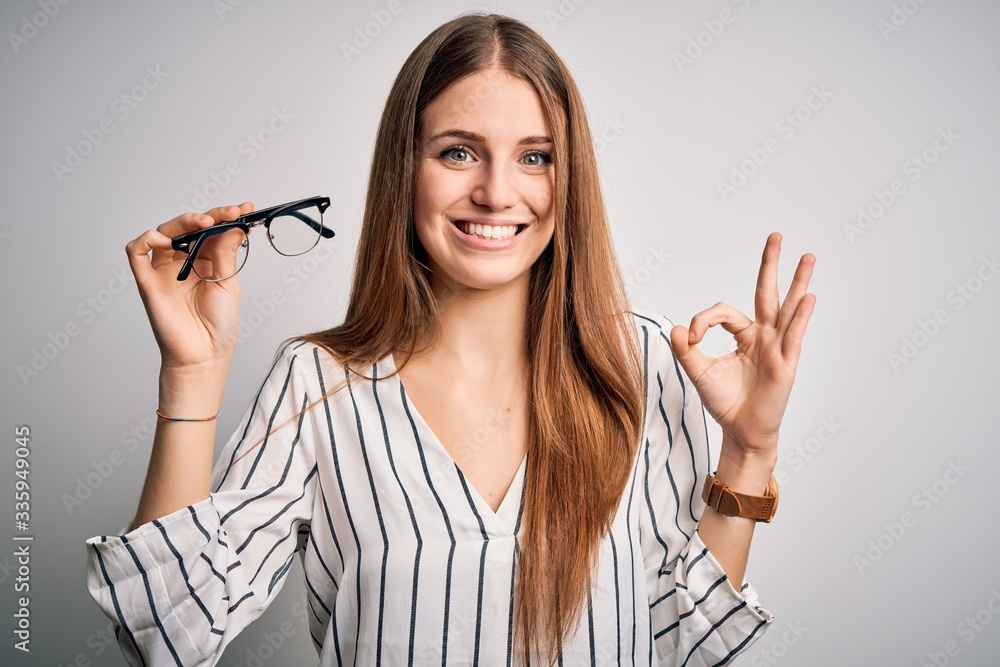 Obraz premium Young beautiful redhead optical woman holding glasses over isolated white background doing ok sign with fingers, excellent symbol