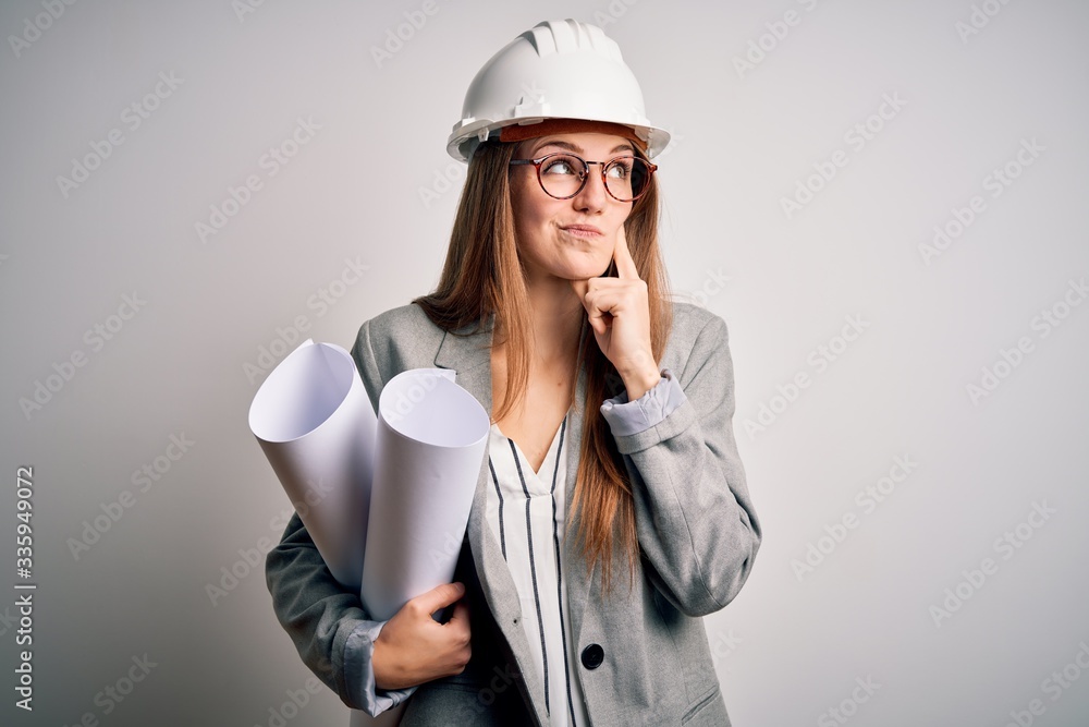 Young beautiful redhead architect woman wearing glasses and safety helmet holding blueprints serious face thinking about question, very confused idea Young beautiful redhead architect woman wearing glasses and safety helmet holding blueprints serious face thinking about question, very confused idea