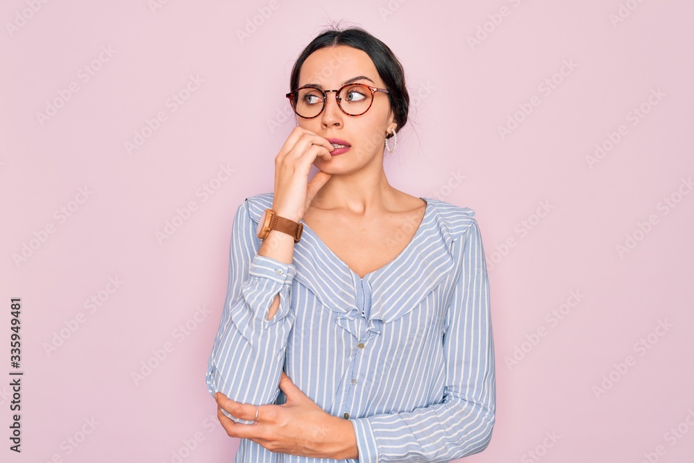 Young beautiful woman wearing casual striped shirt and glasses over pink background looking stressed and nervous with hands on mouth biting nails. Anxiety problem. Young beautiful woman wearing casual striped shirt and glasses over pink background looking stressed and nervous with hands on mouth biting nails. Anxiety problem.