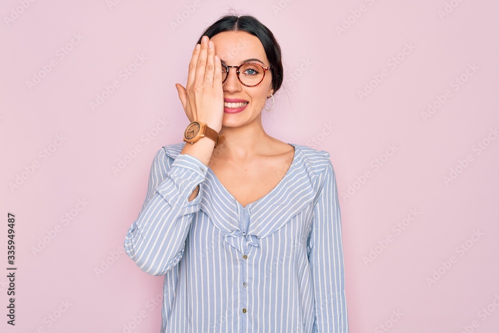 Young beautiful woman wearing casual striped shirt and glasses over pink background covering one eye with hand, confident smile on face and surprise emotion.