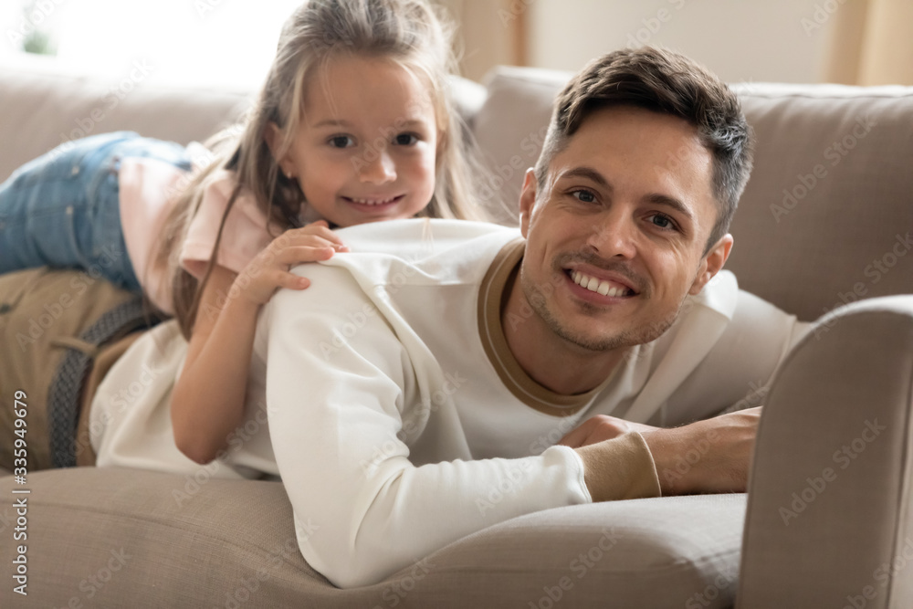 Happy young father lying on comfortable couch with smiling small cute daughter on back. Joyful dad relaxing on sofa with little kid girl, enjoying lazy free weekend time in living room together. Happy young father lying on comfortable couch with smiling small cute daughter on back. Joyful dad relaxing on sofa with little kid girl, enjoying lazy free weekend time in living room together.
