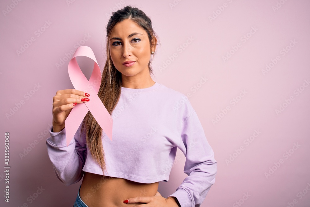 Young beautiful brunette woman holding pink cancer ribbon symbol over isolated background with a confident expression on smart face thinking serious Young beautiful brunette woman holding pink cancer ribbon symbol over isolated background with a confident expression on smart face thinking serious