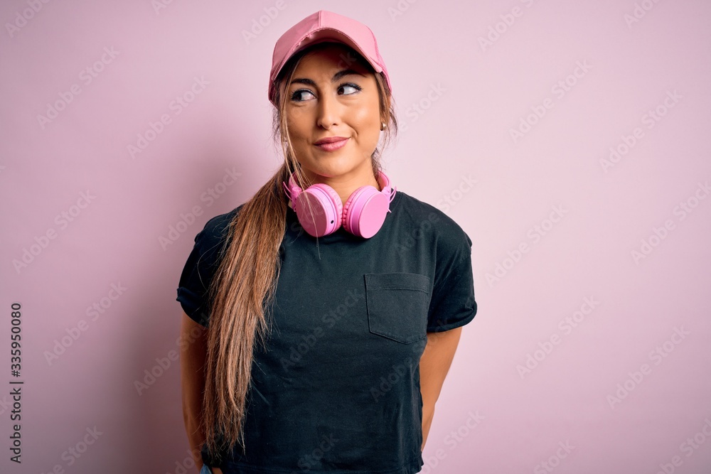Young beautiful fitness sports woman wearing training cap and headphones over pink background smiling looking to the side and staring away thinking. Young beautiful fitness sports woman wearing training cap and headphones over pink background smiling looking to the side and staring away thinking.