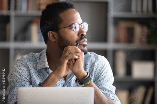 Pensive African American man in glasses distracted from computer work look in distance thinking or pondering, thoughtful biracial male lost in thoughts make plans visualizing, business vision concept photo