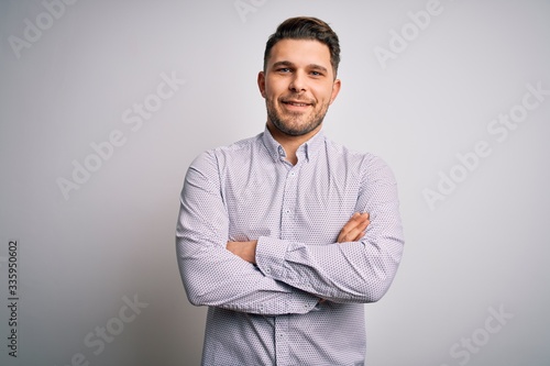 Young business man with blue eyes standing over isolated background happy face smiling with crossed arms looking at the camera. Positive person.