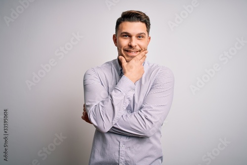 Young business man with blue eyes standing over isolated background looking confident at the camera smiling with crossed arms and hand raised on chin. Thinking positive.