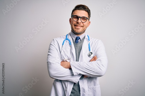 Young doctor man with blue eyes wearing medical coat and stethoscope over isolated background happy face smiling with crossed arms looking at the camera. Positive person.