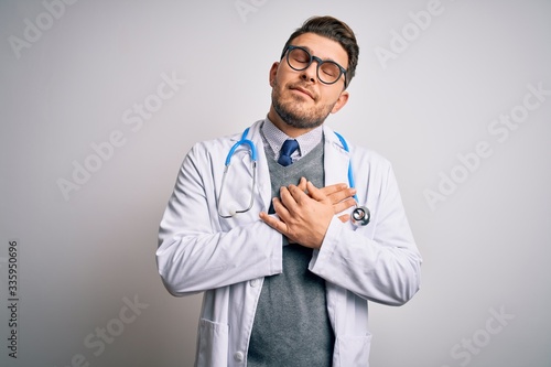 Young doctor man with blue eyes wearing medical coat and stethoscope over isolated background smiling with hands on chest with closed eyes and grateful gesture on face. Health concept.