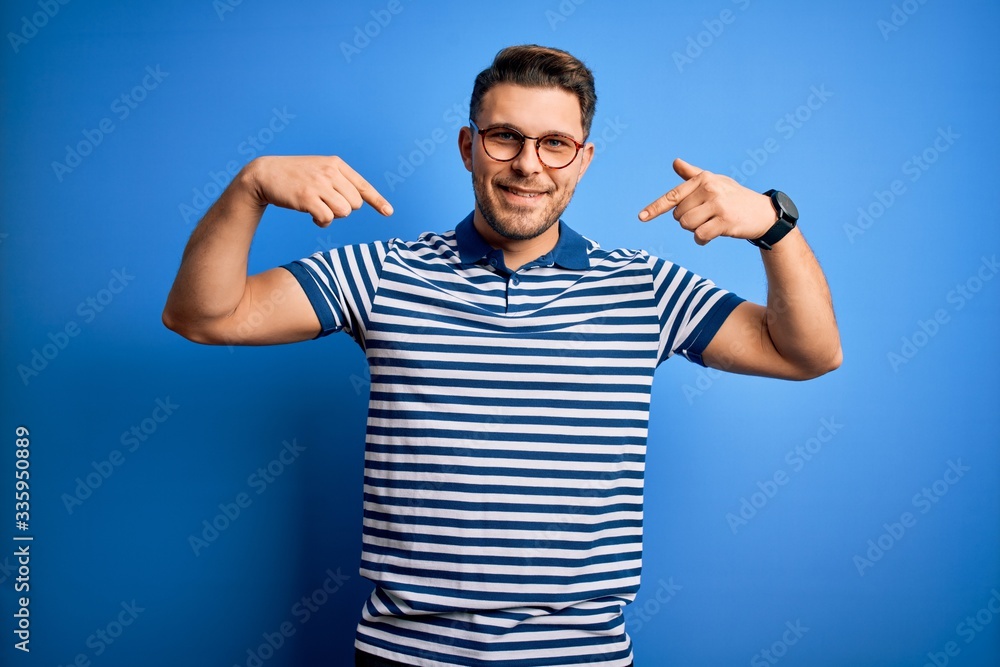 Fototapeta premium Young man with blue eyes wearing glasses and casual striped t-shirt over blue background looking confident with smile on face, pointing oneself with fingers proud and happy.