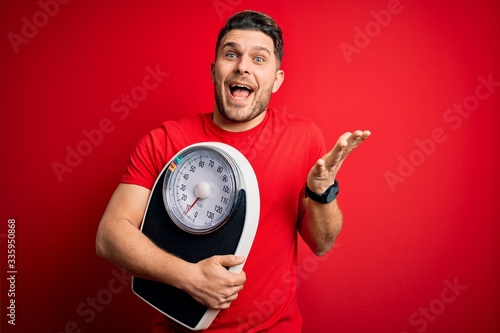 Young fitness man with blue eyes holding scale dieting for healthy weight over red background very happy and excited, winner expression celebrating victory screaming with big smile and raised hands