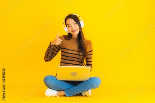 Beautiful portrait young asian woman sit on the floor with laptop and headphone on yellow background
