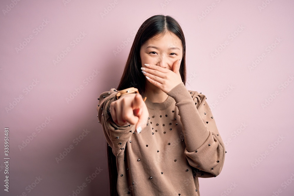 Young beautiful asian woman wearing fashion and elegant sweater over pink solated background laughing at you, pointing finger to the camera with hand over mouth, shame expression Young beautiful asian woman wearing fashion and elegant sweater over pink solated background laughing at you, pointing finger to the camera with hand over mouth, shame expression