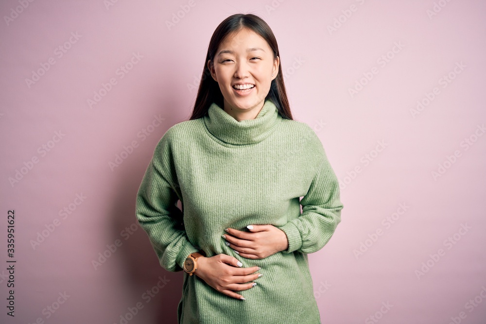 Young beautiful asian woman wearing green winter sweater over pink solated background smiling and laughing hard out loud because funny crazy joke with hands on body. Young beautiful asian woman wearing green winter sweater over pink solated background smiling and laughing hard out loud because funny crazy joke with hands on body.