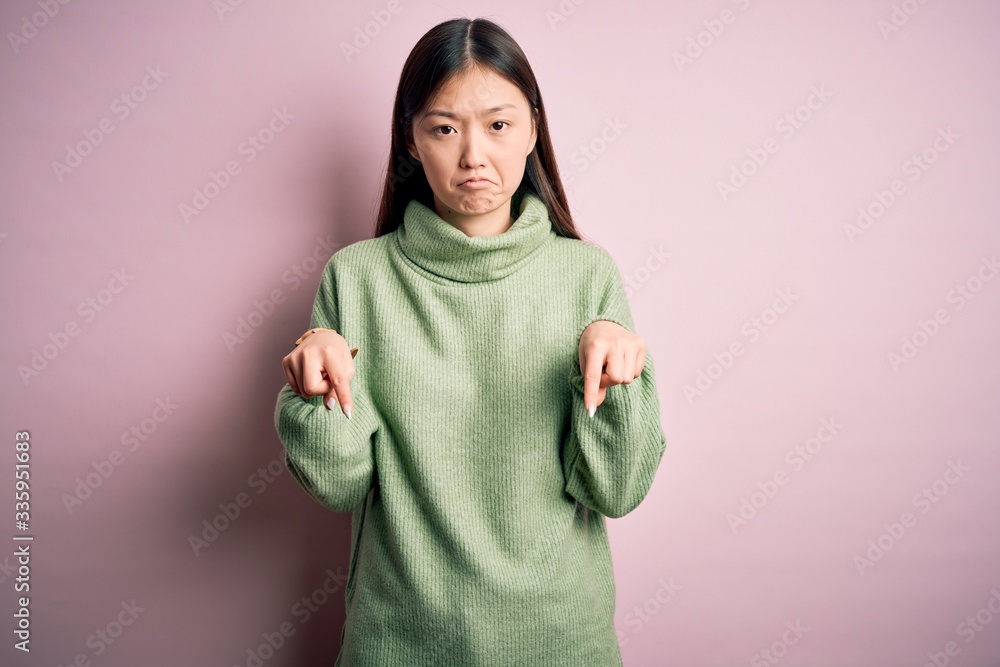 Young beautiful asian woman wearing green winter sweater over pink solated background Pointing down looking sad and upset, indicating direction with fingers, unhappy and depressed. Young beautiful asian woman wearing green winter sweater over pink solated background Pointing down looking sad and upset, indicating direction with fingers, unhappy and depressed.