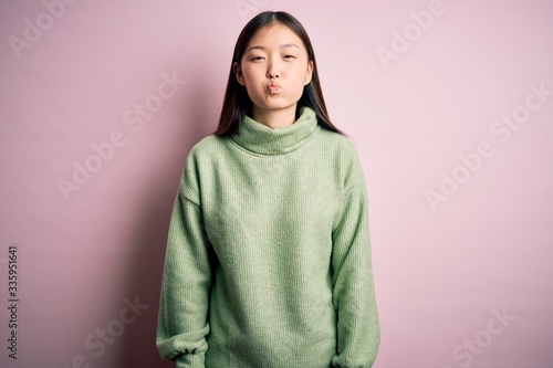 Young beautiful asian woman wearing green winter sweater over pink solated background puffing cheeks with funny face. Mouth inflated with air, crazy expression. © Krakenimages.com