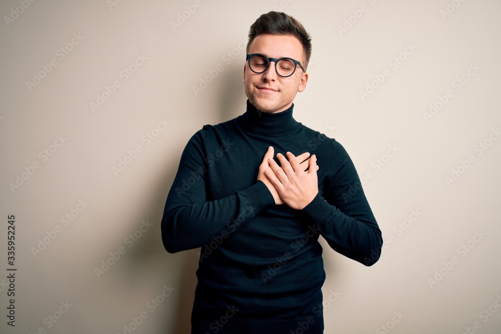 Young handsome caucasian man wearing glasses and casual sweater over isolated background smiling with hands on chest with closed eyes and grateful gesture on face. Health concept. Young handsome caucasian man wearing glasses and casual sweater over isolated background smiling with hands on chest with closed eyes and grateful gesture on face. Health concept.