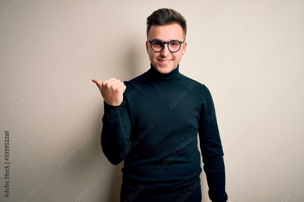 Young handsome caucasian man wearing glasses and casual sweater over isolated background smiling with happy face looking and pointing to the side with thumb up. Young handsome caucasian man wearing glasses and casual sweater over isolated background smiling with happy face looking and pointing to the side with thumb up.