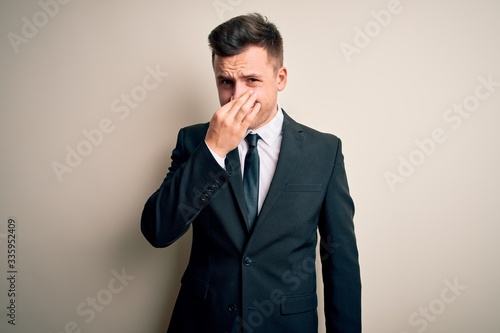 Young handsome business man wearing elegant suit and tie over isolated background smelling something stinky and disgusting, intolerable smell, holding breath with fingers on nose. Bad smell