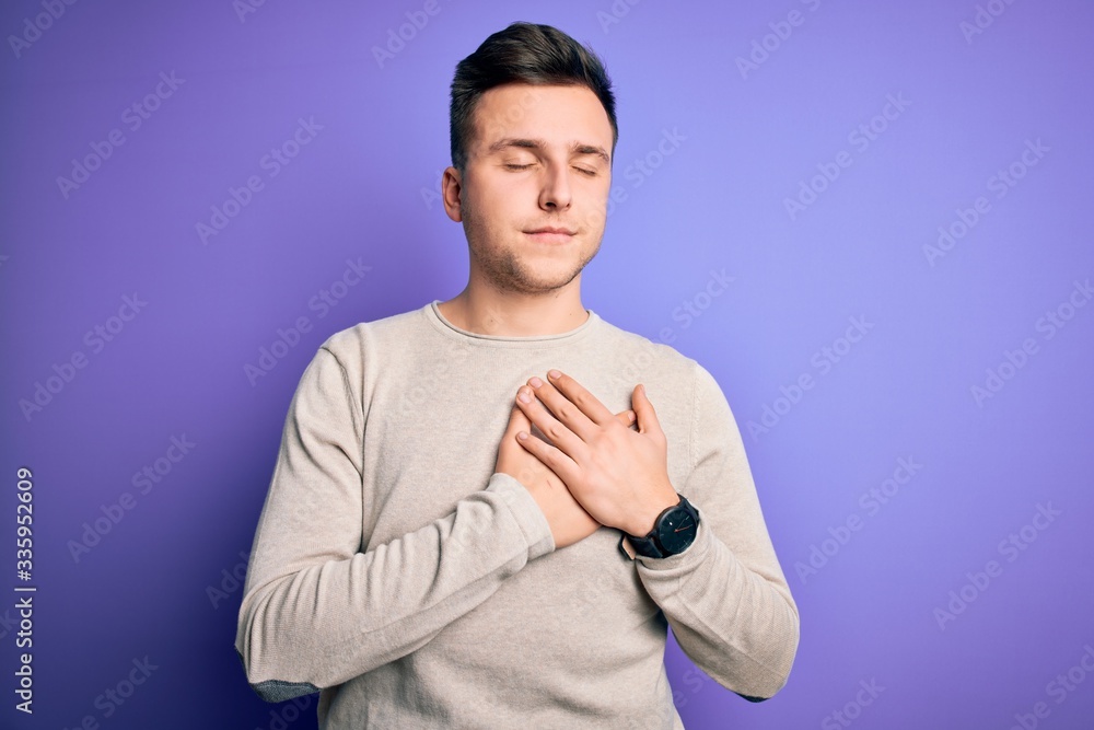 Young handsome caucasian man wearing casual sweater over purple isolated background smiling with hands on chest with closed eyes and grateful gesture on face. Health concept.