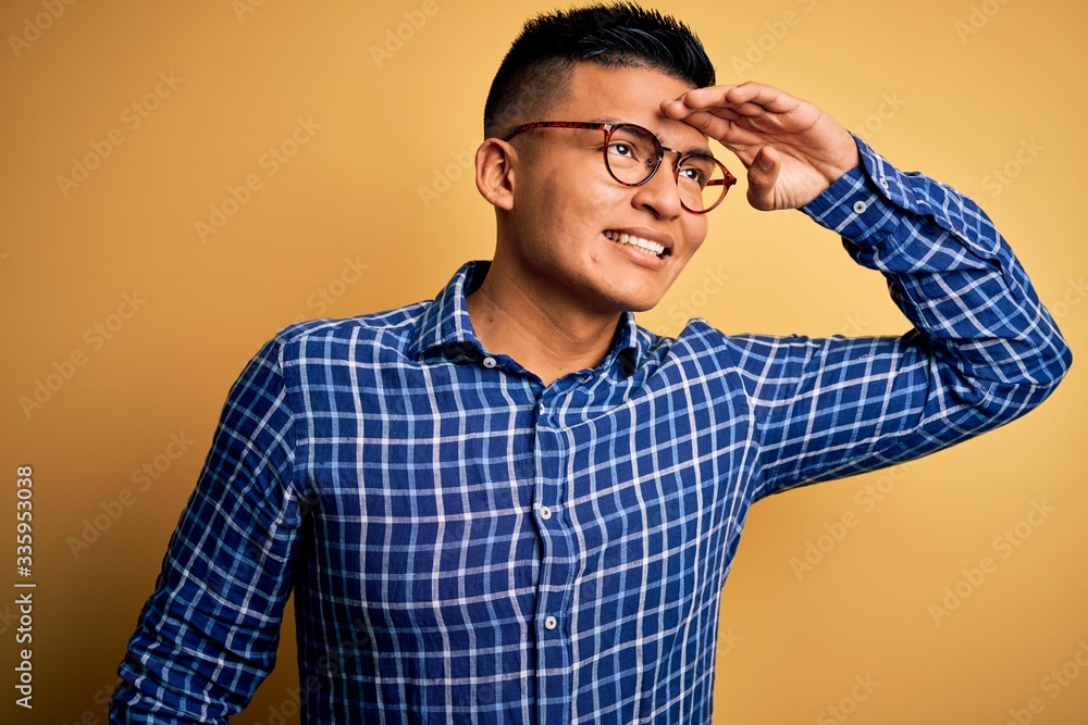 Young handsome latin man wearing casual shirt and glasses over yellow background very happy and smiling looking far away with hand over head. Searching concept. Young handsome latin man wearing casual shirt and glasses over yellow background very happy and smiling looking far away with hand over head. Searching concept.