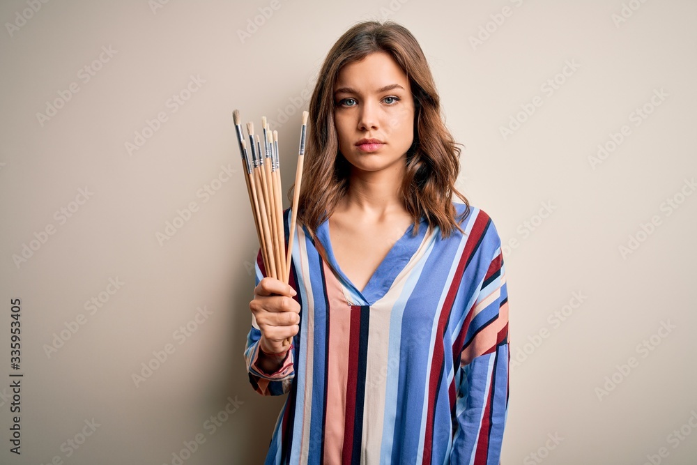 Young blonde painter girl holding drawing brushes over isolated background with a confident expression on smart face thinking serious Young blonde painter girl holding drawing brushes over isolated background with a confident expression on smart face thinking serious