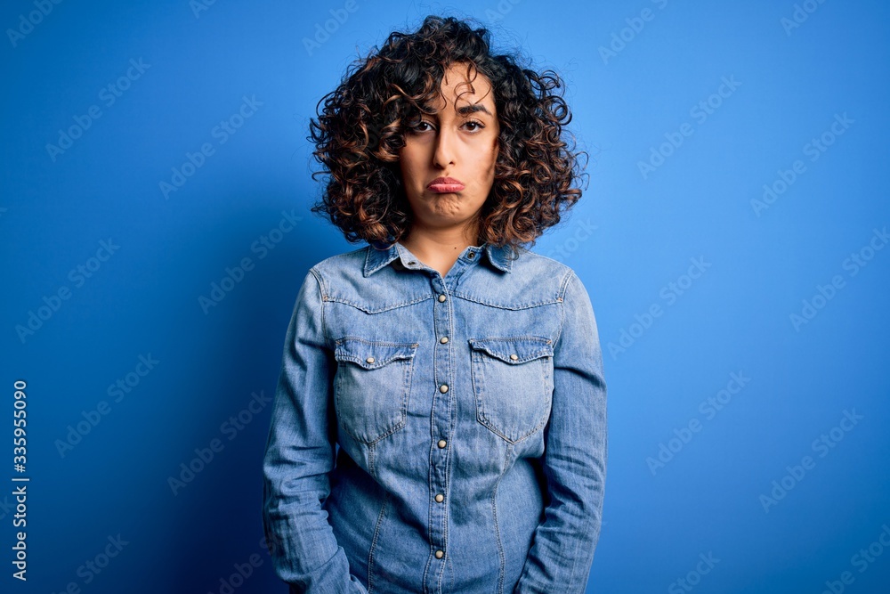 Young beautiful curly arab woman wearing casual denim shirt standing over blue background depressed and worry for distress, crying angry and afraid. Sad expression. Young beautiful curly arab woman wearing casual denim shirt standing over blue background depressed and worry for distress, crying angry and afraid. Sad expression.