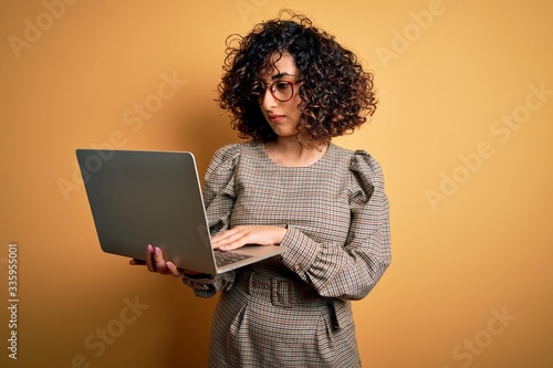 Beautiful arab businesswoman wearing glasses working using laptop over yellow background with a confident expression on smart face thinking serious © Krakenimages.com