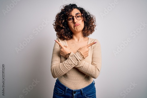 Young beautiful curly arab woman wearing casual t-shirt and glasses over white background Pointing to both sides with fingers, different direction disagree