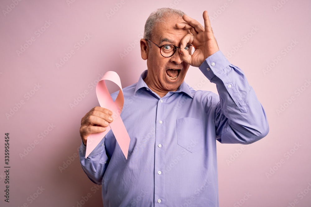 Middle age hoary man holding pink cancer ribbon symbol over isolated background with happy face smiling doing ok sign with hand on eye looking through fingers Middle age hoary man holding pink cancer ribbon symbol over isolated background with happy face smiling doing ok sign with hand on eye looking through fingers