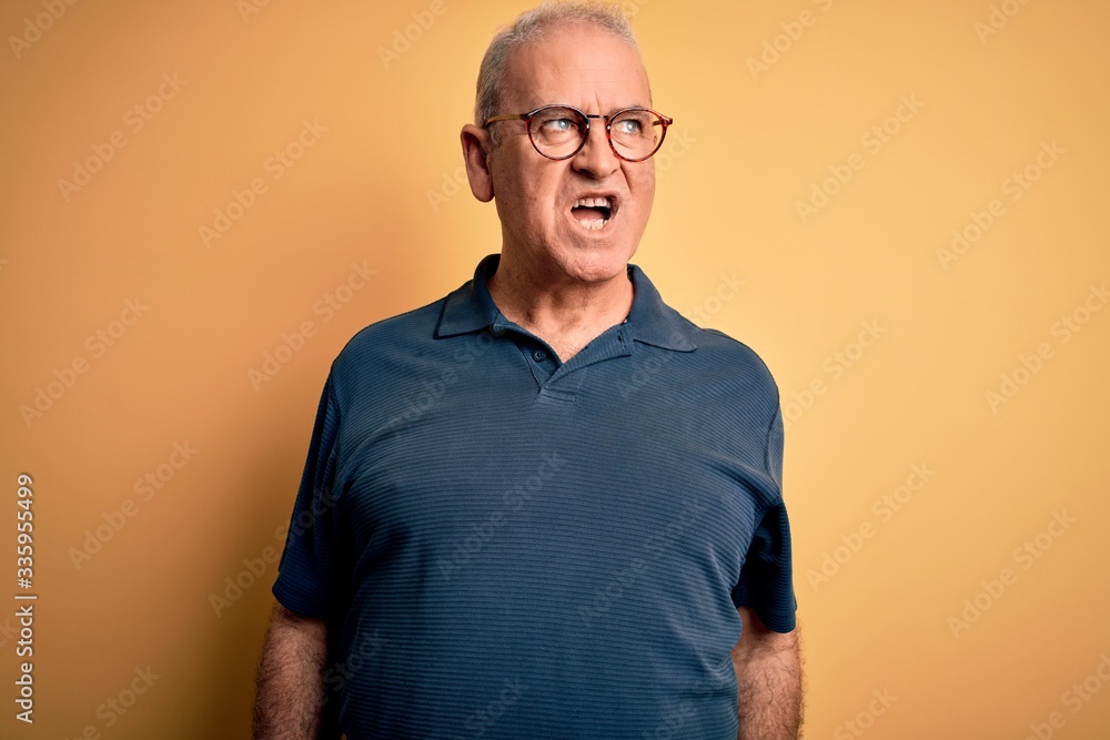 Fototapeta premium Middle age handsome hoary man wearing casual polo and glasses over yellow background angry and mad screaming frustrated and furious, shouting with anger. Rage and aggressive concept.