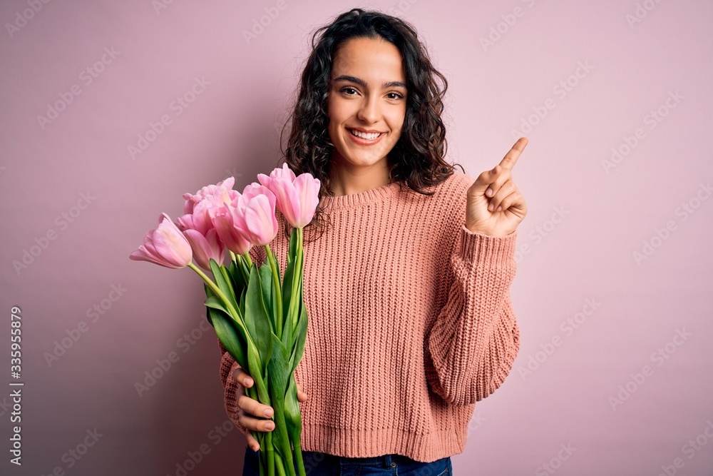 Young beautiful romantic woman with curly hair holding bouquet of pink tulips cheerful with a smile on face pointing with hand and finger up to the side with happy and natural expression Young beautiful romantic woman with curly hair holding bouquet of pink tulips cheerful with a smile on face pointing with hand and finger up to the side with happy and natural expression