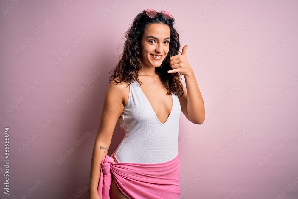 Beautiful woman with curly hair on vacation wearing white swimsuit over pink background smiling doing phone gesture with hand and fingers like talking on the telephone. Communicating concepts. Beautiful woman with curly hair on vacation wearing white swimsuit over pink background smiling doing phone gesture with hand and fingers like talking on the telephone. Communicating concepts.