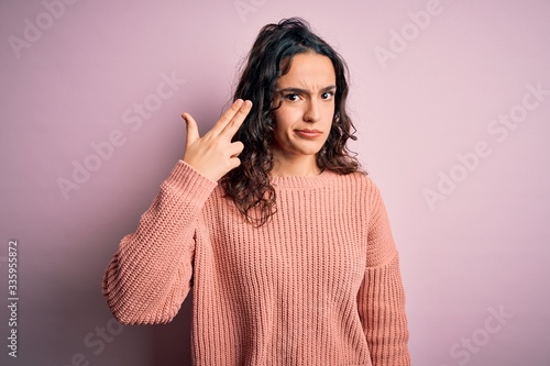 Young beautiful woman with curly hair wearing casual sweater over isolated pink background Shooting and killing oneself pointing hand and fingers to head like gun, suicide gesture. © Krakenimages.com