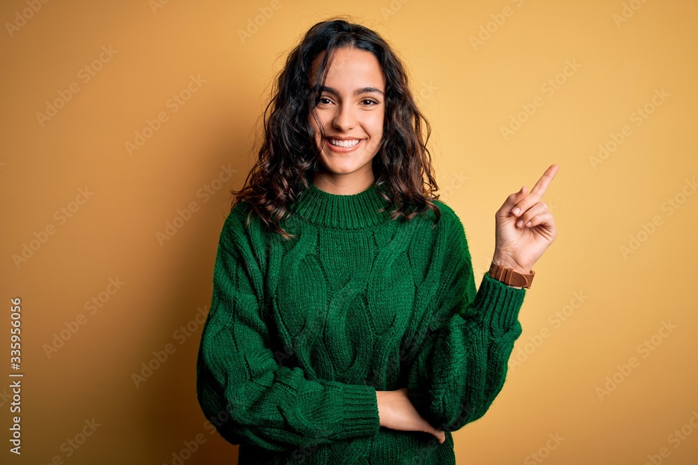 Young beautiful woman with curly hair wearing green casual sweater over yellow background with a big smile on face, pointing with hand and finger to the side looking at the camera. Young beautiful woman with curly hair wearing green casual sweater over yellow background with a big smile on face, pointing with hand and finger to the side looking at the camera.