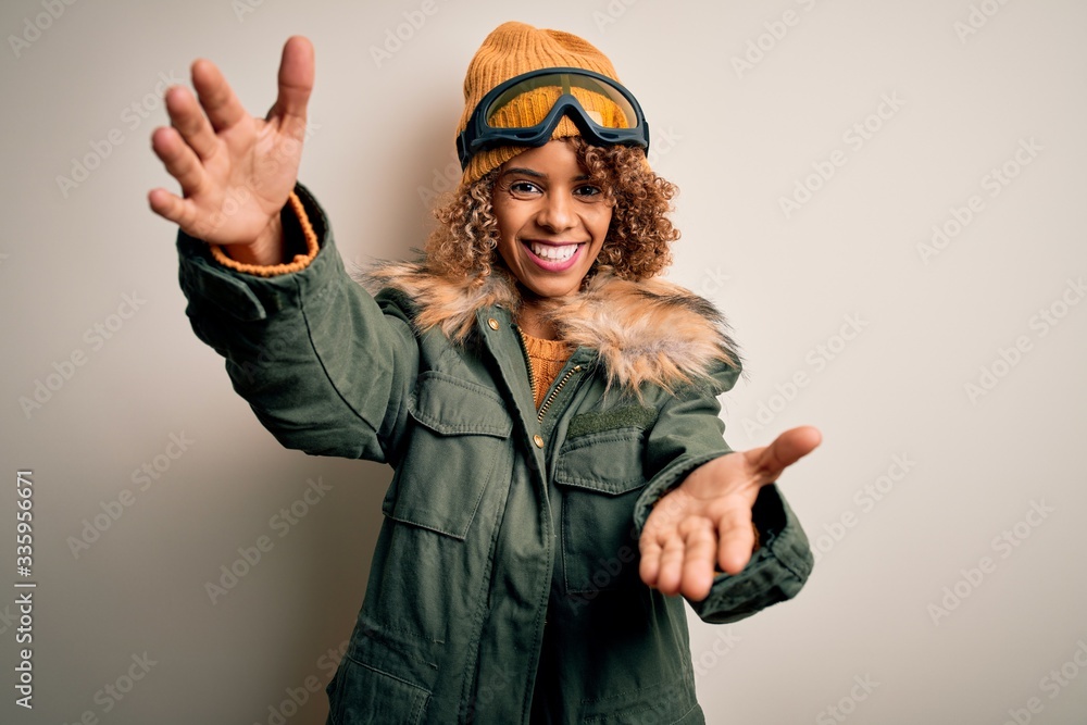 Young african american skier woman with curly hair wearing snow sportswear and ski goggles looking at the camera smiling with open arms for hug. Cheerful expression embracing happiness. Young african american skier woman with curly hair wearing snow sportswear and ski goggles looking at the camera smiling with open arms for hug. Cheerful expression embracing happiness.